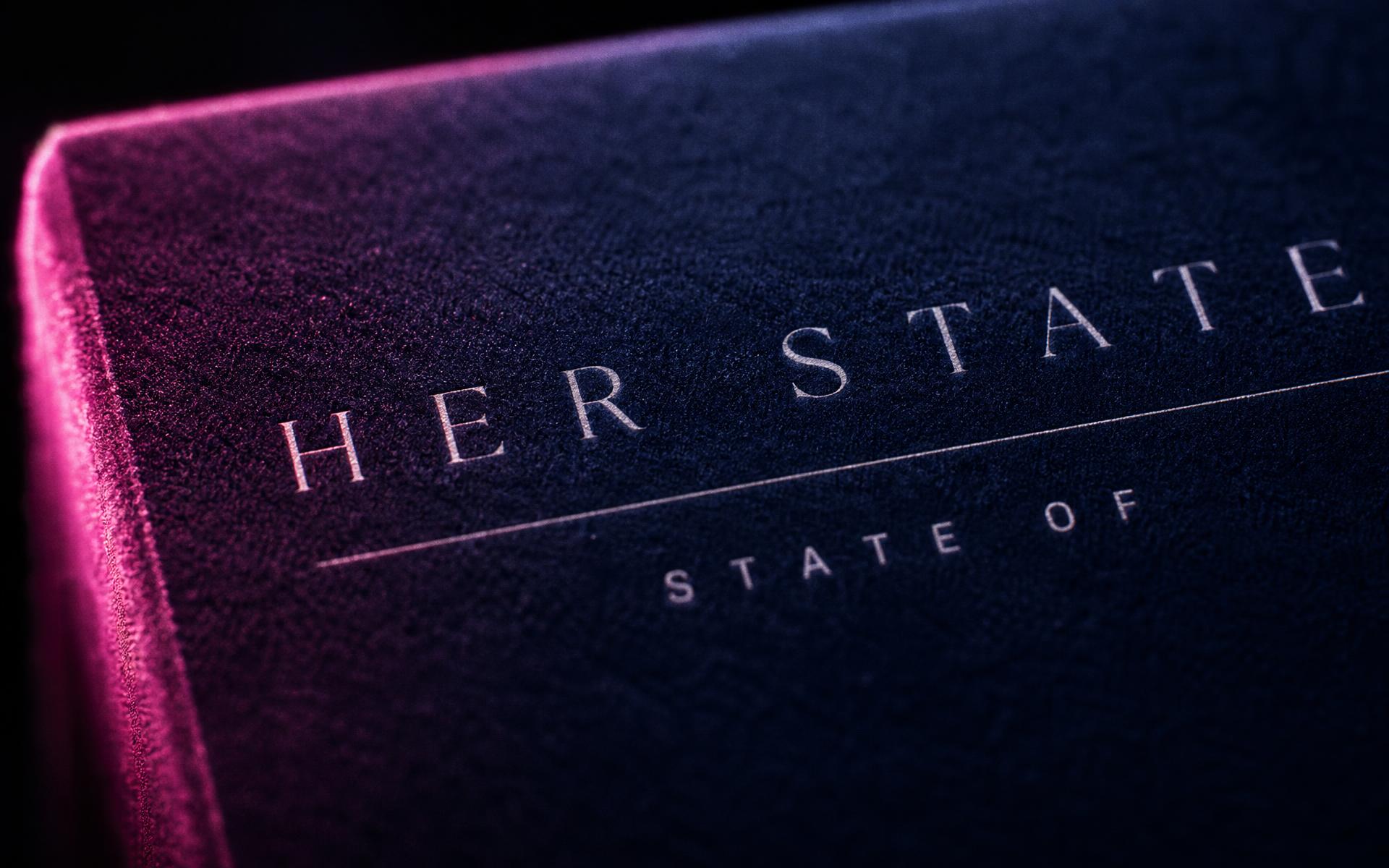 Extreme close-up of the HER STATE lettering printed on the navy velvet package, raked by pink neon light